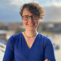 A picture of a woman in a blue dress with glasses and curly hair, smiling at the camera with a background that looks like a balcony overlooking the beach.