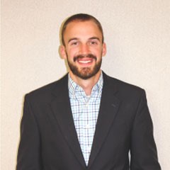 A man in a dark suit with a full, neatly-trimmed beard smiled broadly for the camera with a plain cream background behind him.