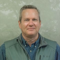 An older man with short-cropped hair and a blue work shirt under a green jacket stands in front of a nondescript background, smiling for the camera.