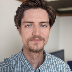 A young man with brown hair brushed back stands in his office. He has a contented expression.